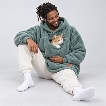 Cyan color: seated man wearing the sherpa pet carrier hoodie with an orange-and-white cat in the front pouch.